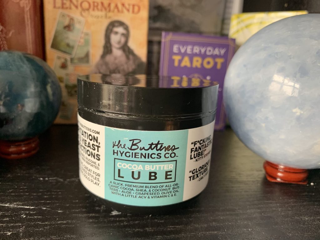 The Butter's Hygienics Co. created several oil-based lubricants. Pictured, is the Cocoa Butter Lube in a black jar atop a table among crystals and Tarot.